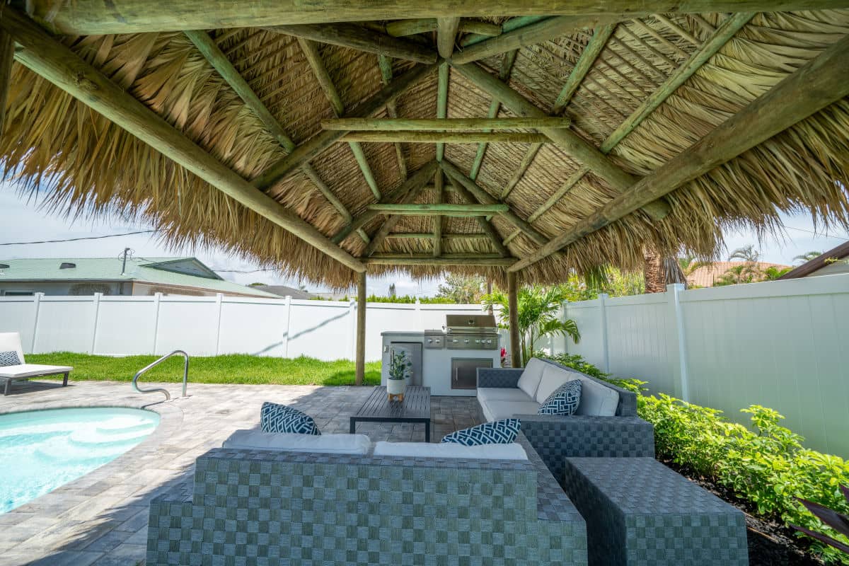 Poolside tiki built over pavers.  Exterior furniture makes a relaxing area next to an outdoor kitchen with a grill under the tiki roof.